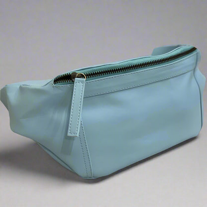 A blue vegan leather sling bag with a zippered front pocket and an adjustable strap, displayed on a textured background.